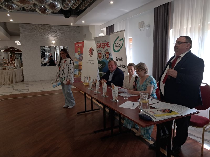Zdjęcie przedstawia jurorów konkursu i laureatkę stojącą koło stołu Jury czytającą nagrodzony wiersz  