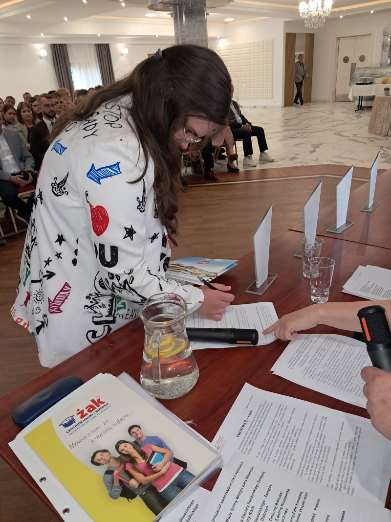 Zdjęcie przedstawia laureatkę konkursu podpisującą na liście odbiór nagrody  