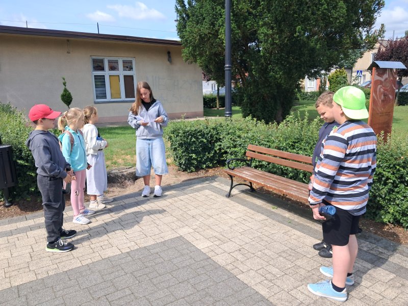 Zdjęcie przedstawia uczestników gry miejskiej odczytujących zadanie z koperty na skępskim Rynku  