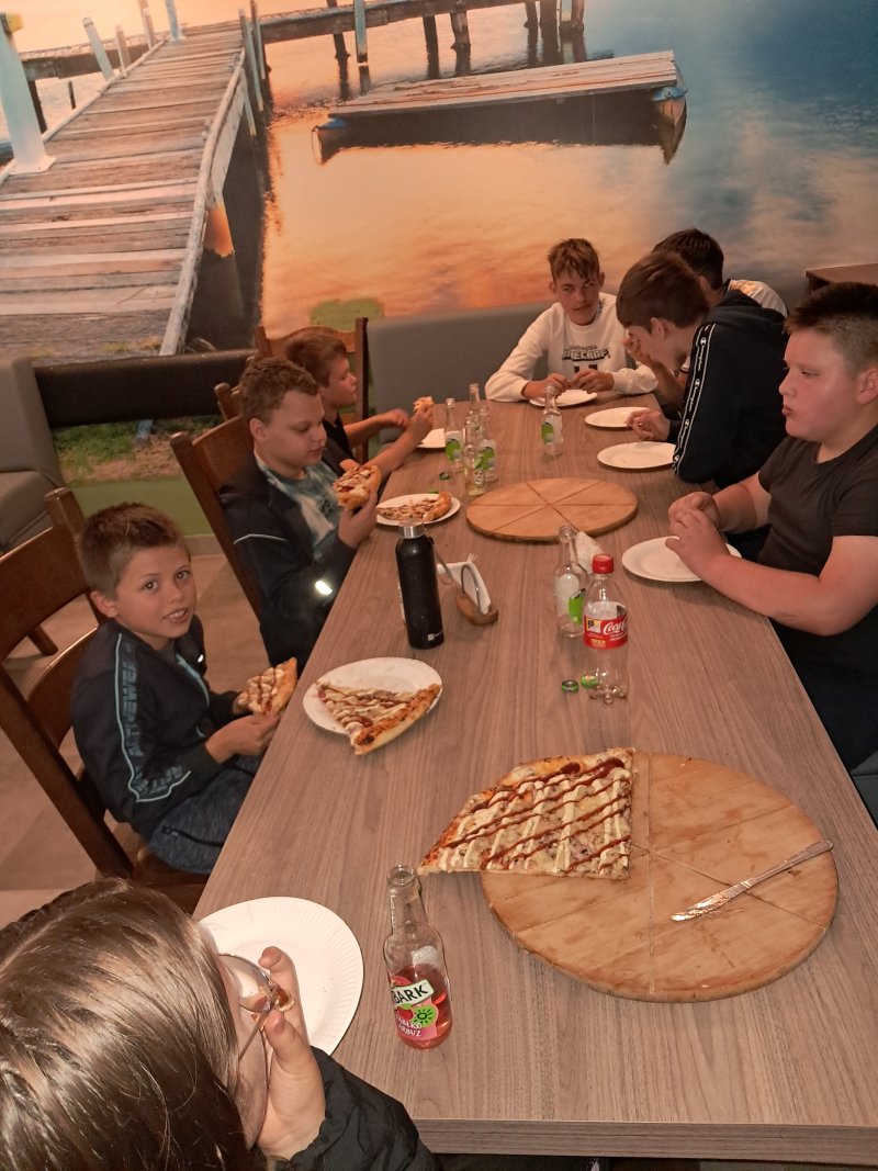 Zdjęcie przedstawia dzieci siedzące przy stoliku w barze jedzące pizzę.  