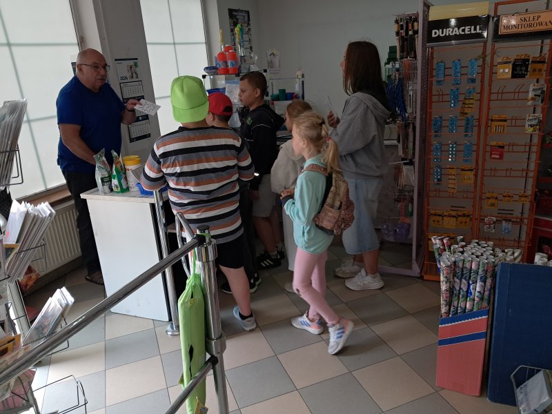 Zdjęcie przedstawia uczestników gry miejskiej odczytujących zadanie z koperty w sklepie papierniczym  