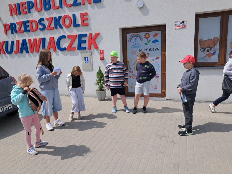 Zdjęcie przedstawia uczestników gry miejskiej odczytujących zadanie z koperty koło przedszkola  