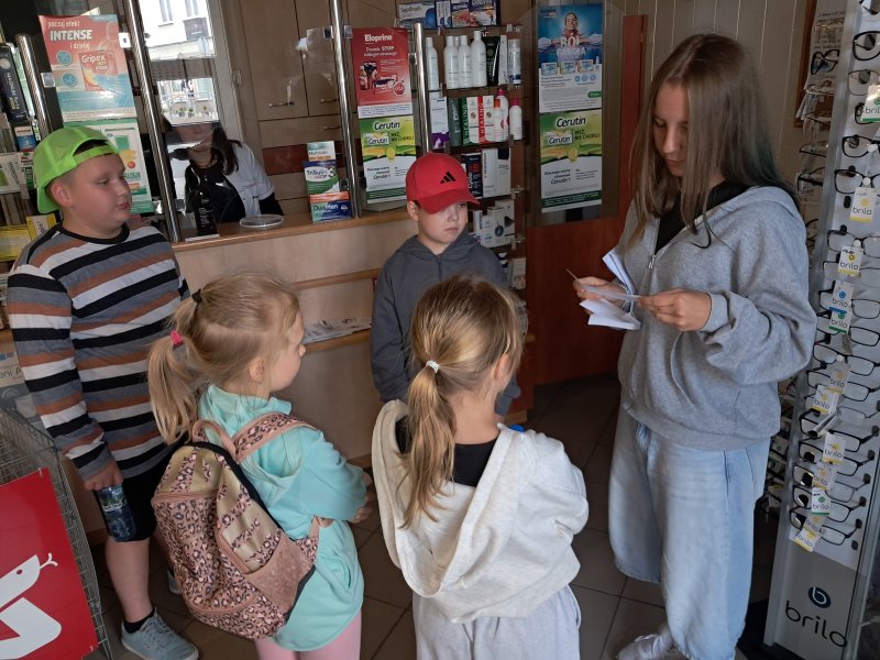 Zdjęcie przedstawia uczestników gry miejskiej odczytujących zadanie z koperty w aptece  