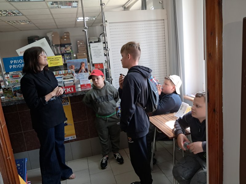 Zdjęcie przedstawia dzieci w trakcie wizyty na poczcie. Dzieci stoją przed pracownikiem, który do nich mówi.  