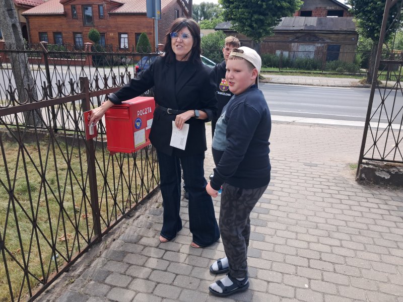 Zdjęcie przedstawia panią stojącą wraz z dziećmi przy skrzynce pocztowej na listy  