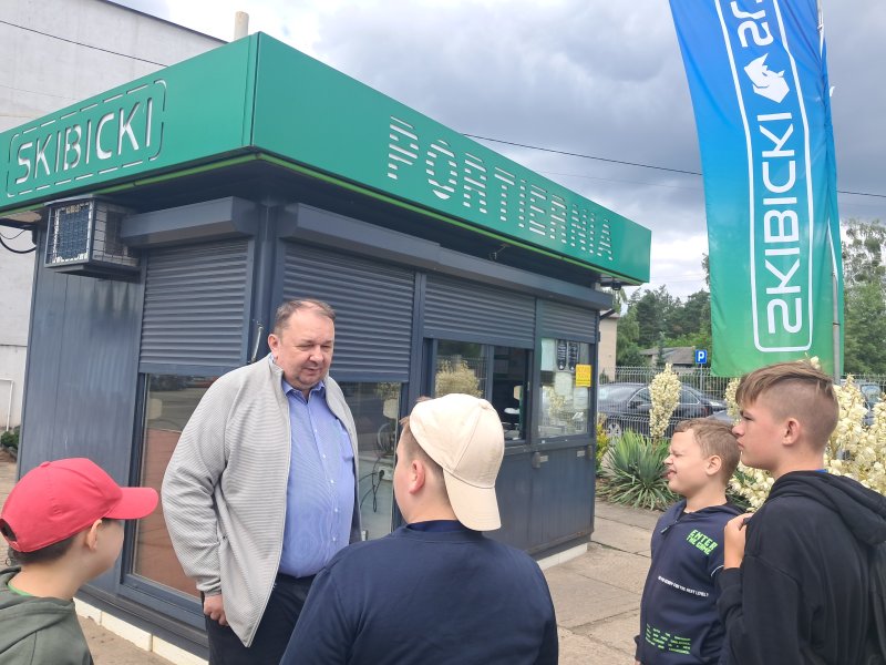Zdjęcie przedstawia dzieci w trakcie wizyty firmie robiącej zabudowy samochodów ciężarowych. Dzieci stoją przed pracownikiem firmy, który do nich mówi.  