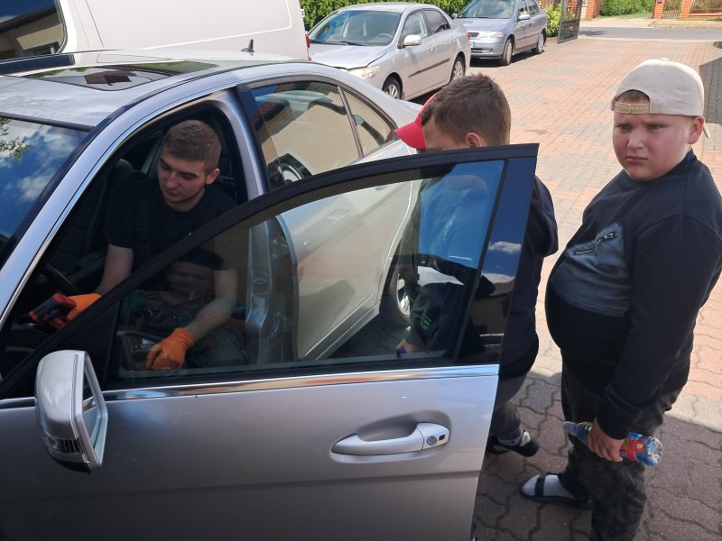 Zdjęcie przedstawia dzieci w trakcie wizyty w warsztacie samochodowym. Na zdjęciu dzieci stoją obok samochodu, w którym siedzi pracownik warsztatu.  