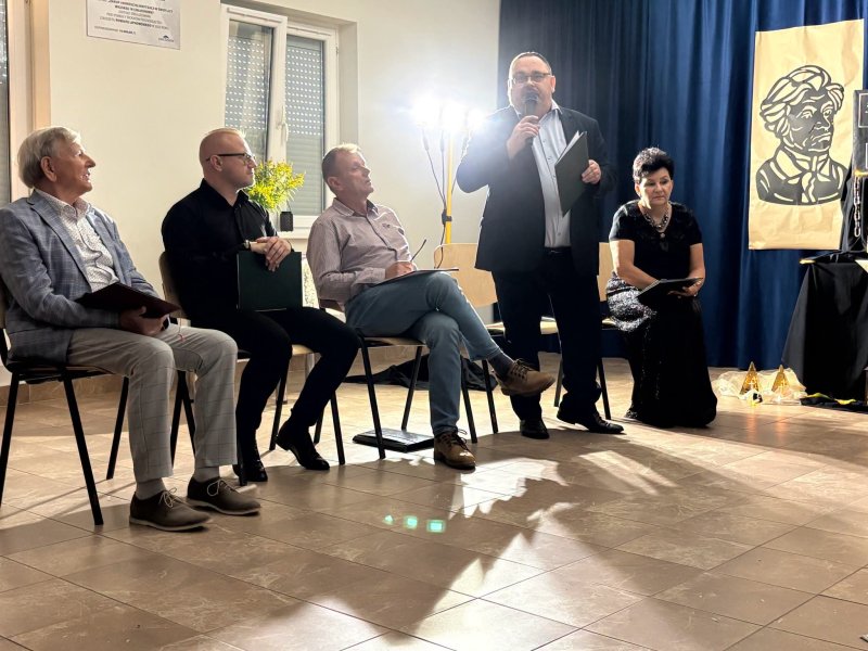 Dyrektor skępskiej biblioteki stoi w świetlicy i przemawia przez mikrofon. Obok na krzesłach siedzą pozostali uczestnicy wydarzenia.  