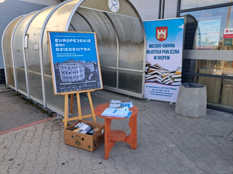 Widok ogólny na wystawiony stolik, roll-up biblioteki i tablicę Europejskich Dni Dziedzictwa wystawione przed sklepem, przed którym odbywa się quiz wiedzy dla mieszkańców  