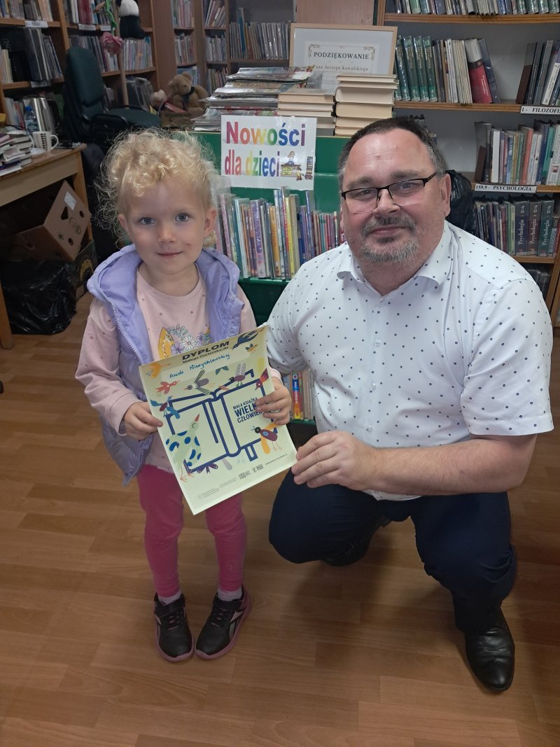Dyrektor Biblioteki kuca przy stojącej dziewczynce, która trzyma w ręku dyplom małego czytelnika i pozują do wspólnej fotografii  